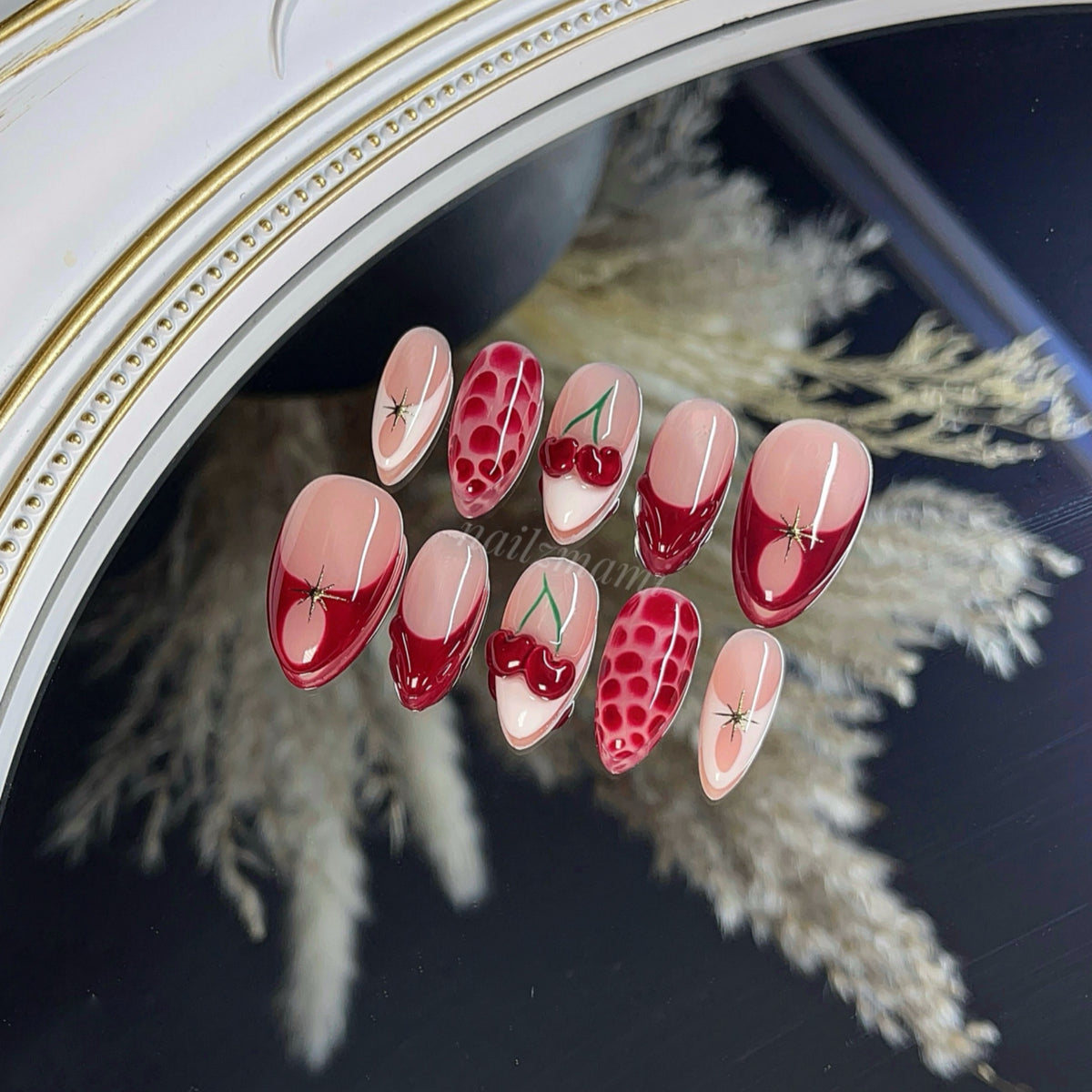 a gel press-on nail set with red french, red 3d cherries, white french, red blooming gel, 3d gel design, and gold chrome in almond shape.