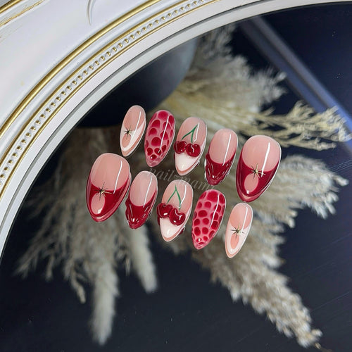 a gel press-on nail set with red french, red 3d cherries, white french, red blooming gel, 3d gel design, and gold chrome in almond shape.
